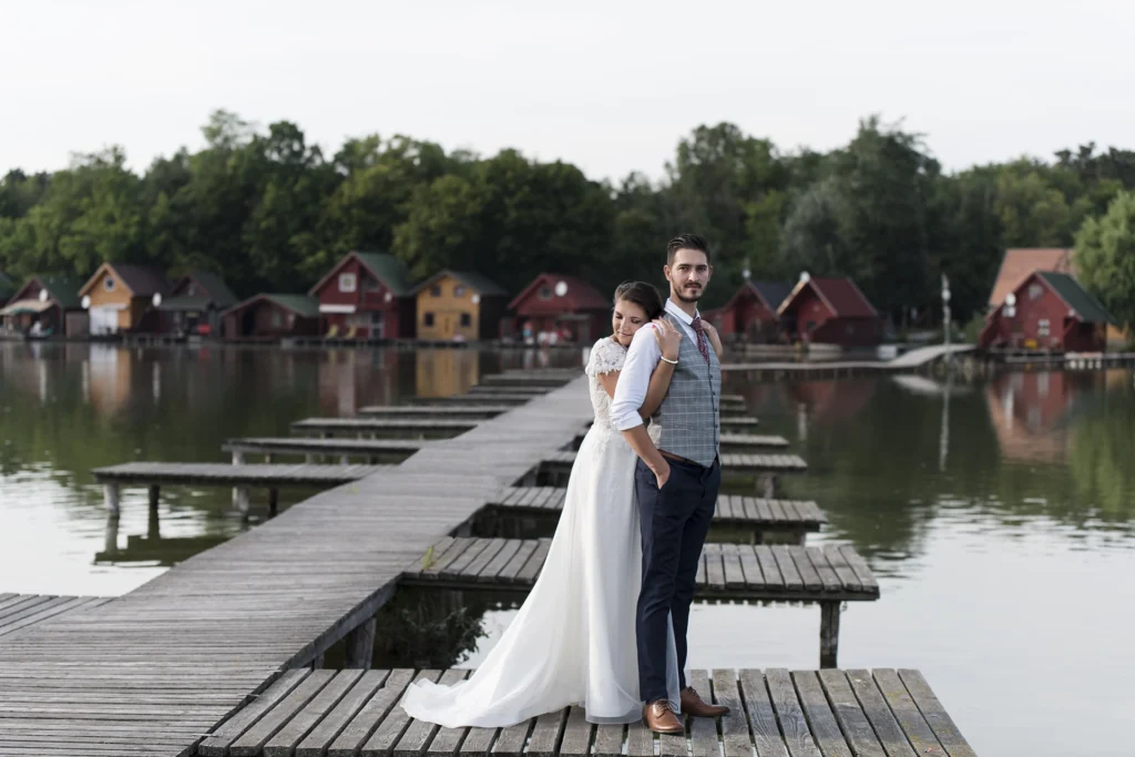 Romantikus esküvői fotó Tatai Öreg-tónál, ahol a menyasszony csukott szemmel, hátulról öleli férjét a stégen állva, meghitt pillanatban