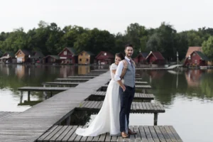 Romantikus esküvői fotó Tatai Öreg-tónál, ahol a menyasszony csukott szemmel, hátulról öleli férjét a stégen állva, meghitt pillanatban