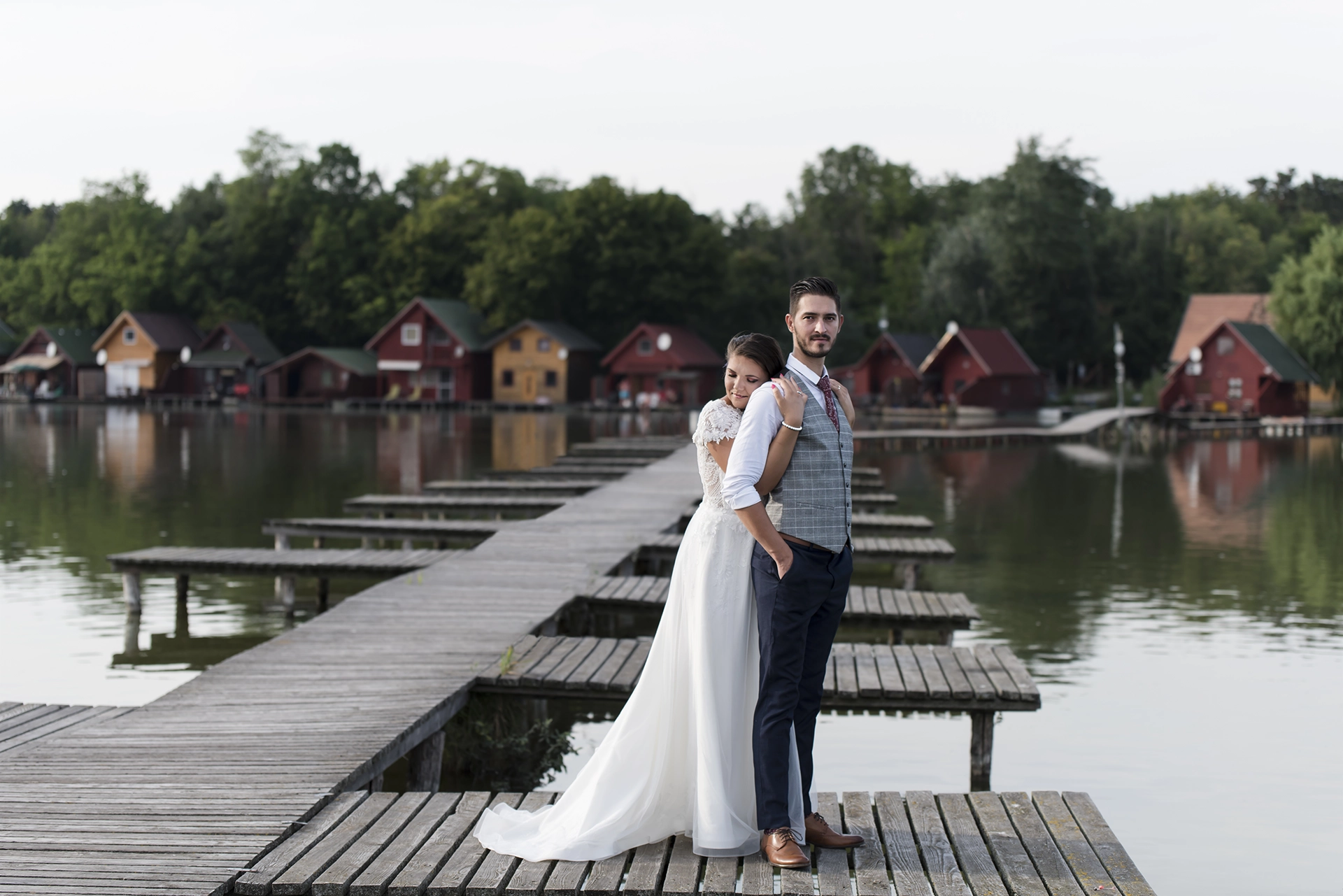 Romantikus esküvői fotó Tatai Öreg-tónál, ahol a menyasszony csukott szemmel, hátulról öleli férjét a stégen állva, meghitt pillanatban