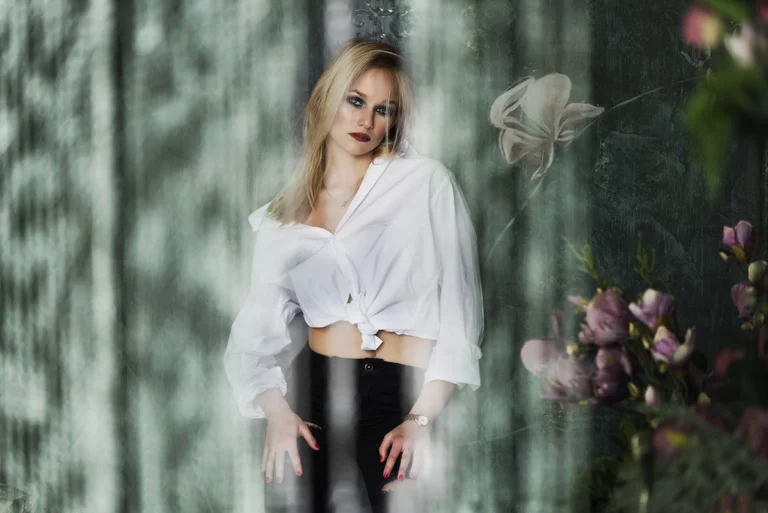 Fehér inget viselő szőke hajú nő pózol a kamerába stúdió háttér előtt, lágy fényben, glamour stílusban