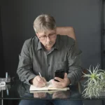 Szürke inget viselő férfi üzletember telefonról jegyzetel az irodában