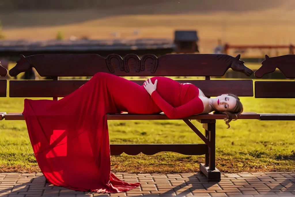 Kezdőlap Kismama piros ruhában fekszik aranyórában a padon, lovas parkban kültéri fotózás